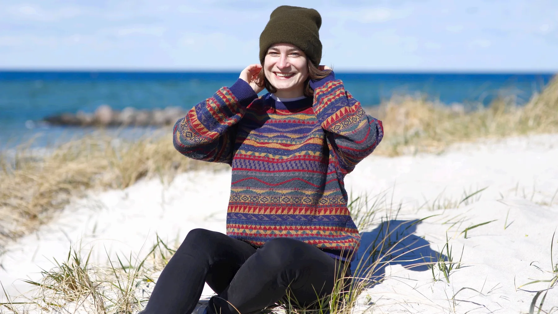 Lachende Frau mit Mütze und gemustertem Pullover in den Dünen, Meer im Hintergrund